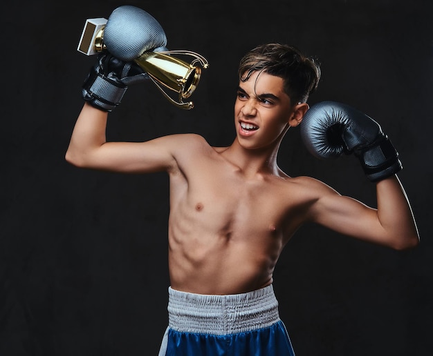 Un alegre joven campeón de boxeador sin camisa con guantes sostiene una copa de ganador. Aislado en un fondo oscuro.