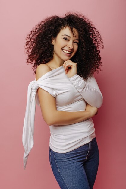 Alegre joven caucásica con cabello rizado negro sonríe blanco como la nieve a la cámara sobre fondo rosa Chica viste blusa blanca y jeans Concepto de emociones de personas de belleza