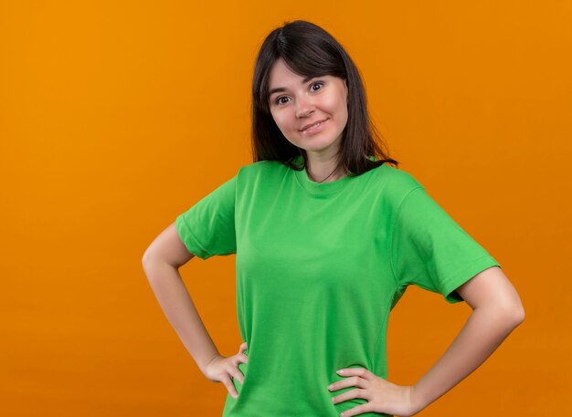 Alegre joven caucásica en camisa verde pone las manos en la cintura y mira a la cámara sobre fondo naranja aislado con espacio de copia