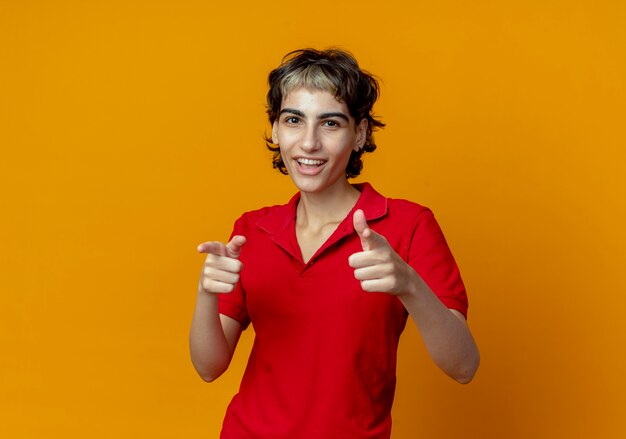 Alegre joven caucásica con corte de pelo pixie apuntando a la cámara aislada sobre fondo naranja con espacio de copia