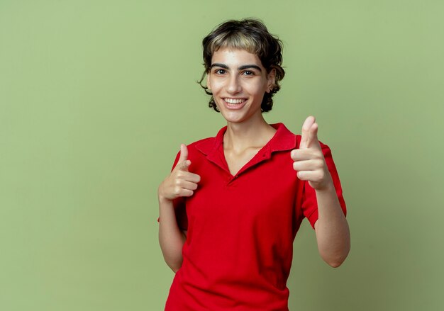 Alegre joven caucásica con corte de pelo pixie apuntando a la cámara aislada sobre fondo verde oliva con espacio de copia