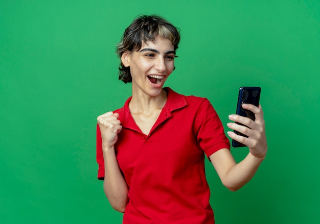 Foto gratuita alegre joven caucásica con corte de pelo pixie sosteniendo y mirando el teléfono móvil con el puño cerrado