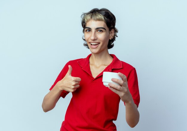 Alegre joven caucásica con corte de pelo pixie sosteniendo la taza y mostrando el pulgar hacia arriba aislado sobre fondo blanco con espacio de copia