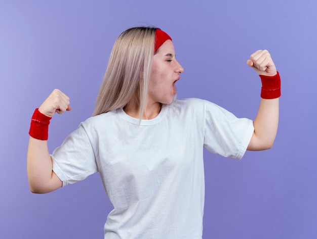 Alegre joven caucásica deportiva con tirantes vistiendo diadema y muñequeras tensa bíceps