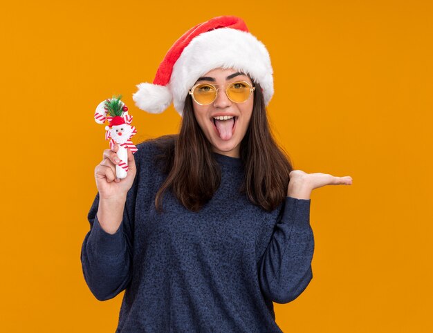 Alegre joven caucásica en gafas de sol con gorro de Papá Noel saca la lengua y sostiene bastón de caramelo aislado en la pared naranja con espacio de copia