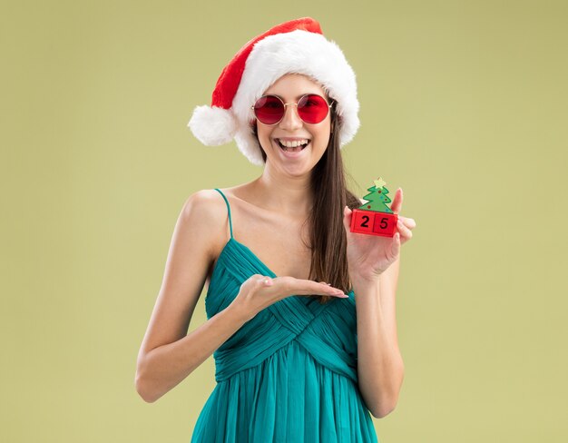 Alegre joven caucásica en gafas de sol con sombrero de santa sosteniendo y apuntando al adorno del árbol de Navidad con la mano