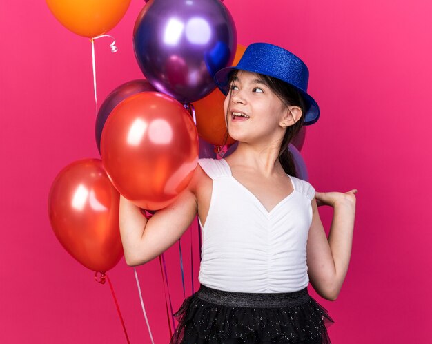 Alegre joven caucásica con gorro de fiesta azul de pie delante de globos de helio mirando aislado en la pared rosa con espacio de copia