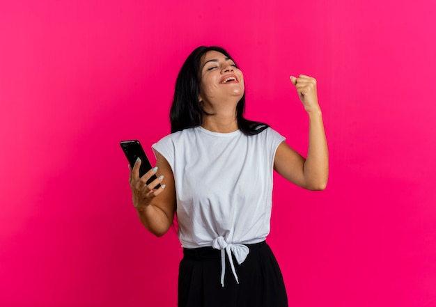 Alegre joven caucásica mantiene el puño y sostiene el teléfono de pie con los ojos cerrados