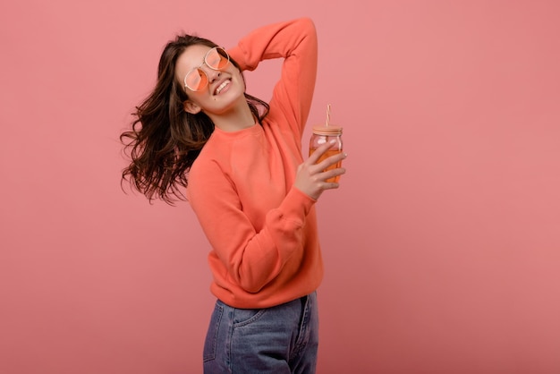 Alegre joven caucásica morena con gafas posa alegremente con bebida en las manos sobre fondo rosa Belleza personas emociones y concepto de vacaciones