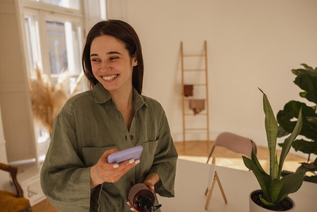 Alegre joven caucásica sosteniendo un teléfono inteligente moderno en sus manos mirando hacia otro lado mientras está de pie en la habitación Morena con una sonrisa en su rostro lee buenas noticias Dispositivo de tecnología y concepto de emociones