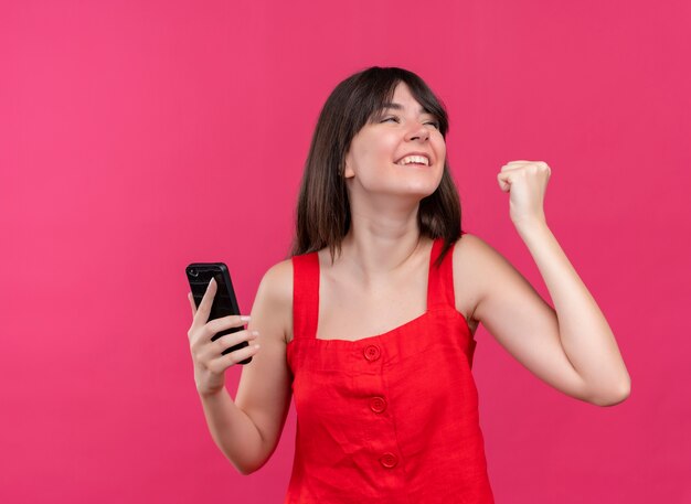 Alegre joven caucásica sosteniendo el teléfono y sosteniendo el puño mirando hacia el lado sobre fondo rosa aislado con espacio de copia