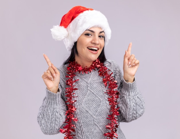 Alegre joven caucásica vistiendo gorro de navidad y guirnalda de oropel alrededor del cuello apuntando hacia arriba aislado en la pared blanca