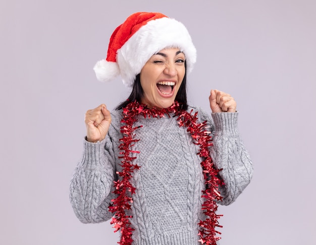 Alegre joven caucásica vistiendo gorro de navidad y guirnalda de oropel alrededor del cuello mirando a la cámara guiñando un ojo haciendo sí gesto aislado sobre fondo blanco.