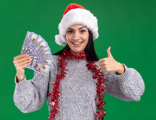 Alegre joven caucásica vistiendo gorro de navidad y guirnalda de oropel alrededor del cuello sosteniendo dinero mostrando el pulgar hacia arriba aislado en la pared verde