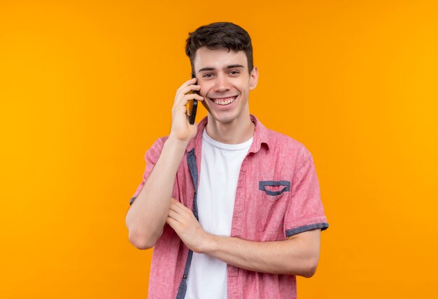 Alegre joven caucásico con camisa rosa habla por teléfono en la pared naranja aislada