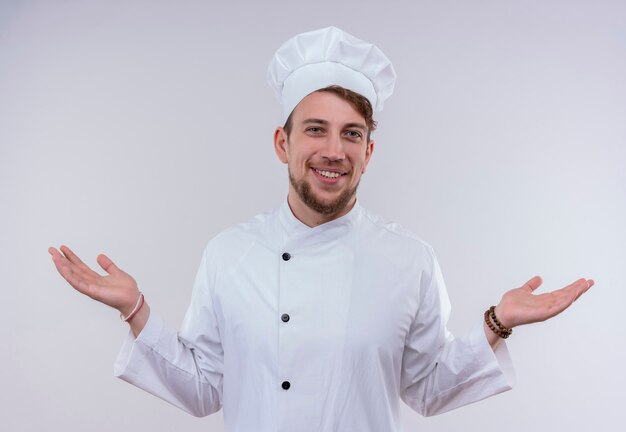 Un alegre joven chef barbudo hombre vestido con uniforme de cocina blanco y sombrero abriendo sus manos amigablemente mientras mira en una pared blanca