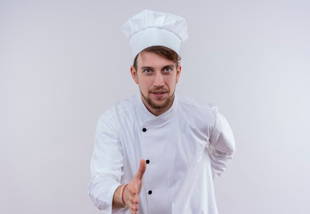 Un alegre joven chef barbudo hombre vestido con uniforme de cocina blanco y sombrero estirando la mano para agitar para recibir a alguien en una pared blanca