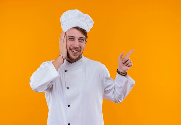 Un alegre joven chef barbudo con uniforme blanco apuntando hacia arriba con el dedo índice mientras sostiene la mano en la cara y mira en una pared naranja