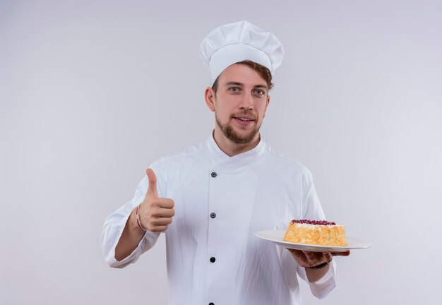 Un alegre joven chef barbudo con uniforme blanco de cocina y sombrero sosteniendo un plato con pastel y mostrando los pulgares hacia arriba mientras mira en una pared blanca