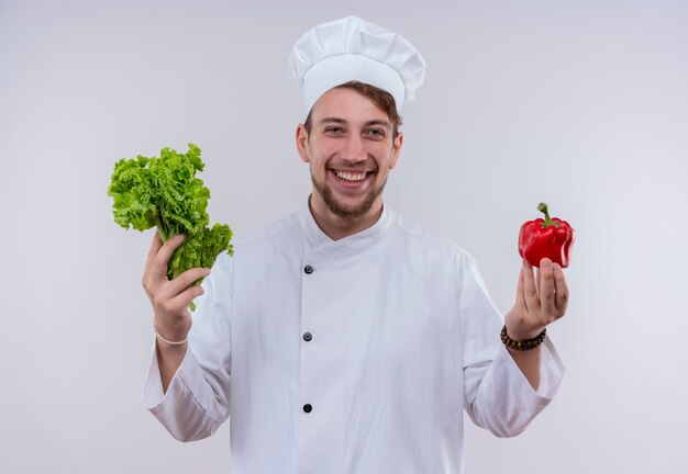 Un alegre joven chef barbudo con uniforme de cocina blanco y sombrero sosteniendo lechuga de hoja verde y pimiento rojo mientras mira en una pared blanca