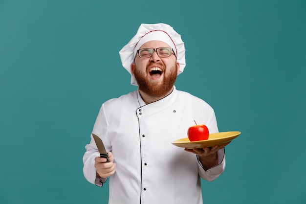 Alegre joven chef masculino con uniforme de anteojos y gorra sosteniendo un plato con manzana y un cuchillo riéndose con los ojos cerrados aislado en el fondo azul