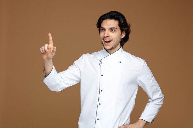 alegre joven chef uniformado manteniendo la mano en la cintura mirando al lado apuntando hacia arriba aislado en fondo marrón