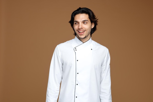 Un alegre joven chef uniformado mirando a un lado sonriendo aislado de fondo marrón