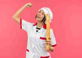 Foto gratuita alegre joven cocinera en uniforme de chef sosteniendo un palito de pan gesticulando fuerte aislado en rosa