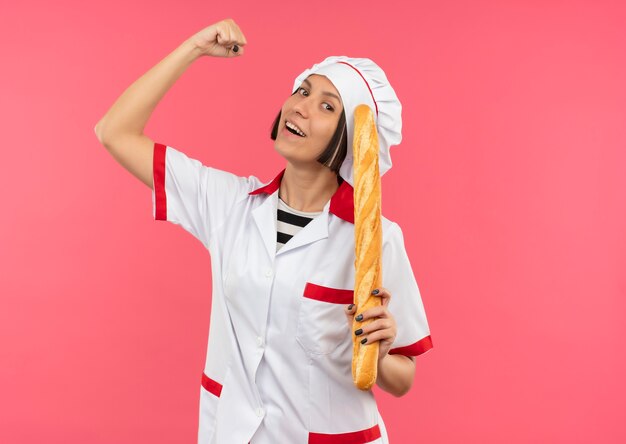 Alegre joven cocinera en uniforme de chef sosteniendo un palito de pan gesticulando fuerte aislado en rosa