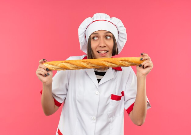 Alegre joven cocinera en uniforme de chef sosteniendo un palito de pan mirando a un lado y mostrando la lengua aislada en rosa