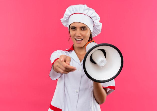 Alegre joven cocinera vistiendo uniforme de chef sosteniendo el altavoz mostrando su gesto en la pared rosa aislada con espacio de copia