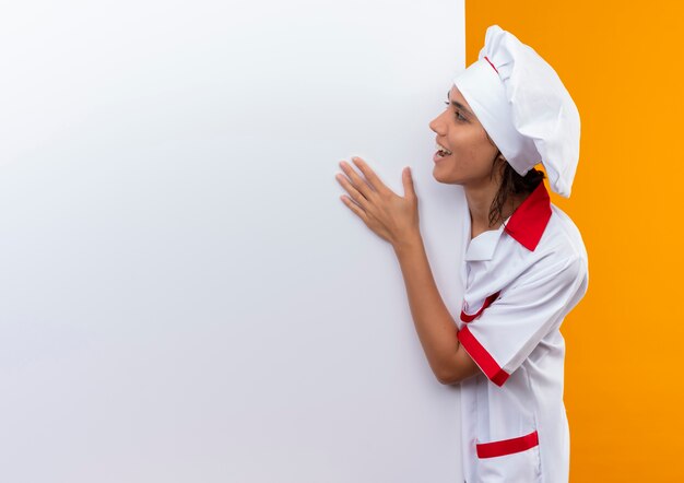 Alegre joven cocinera vistiendo uniforme de chef sosteniendo y mirando la pared blanca en la pared amarilla aislada con espacio de copia