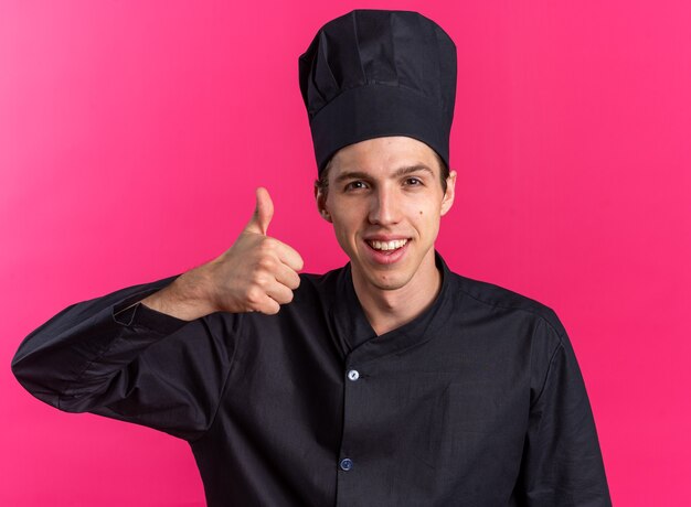 Alegre joven cocinero rubio en uniforme de chef y gorra mostrando el pulgar hacia arriba