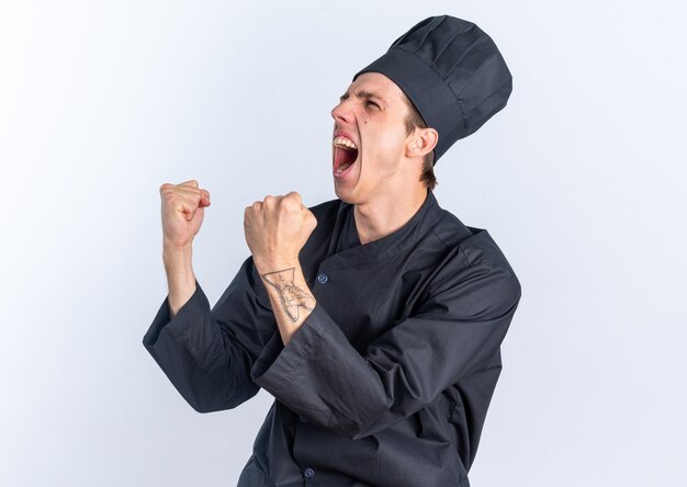 Alegre joven cocinero de sexo masculino rubio en uniforme de chef y gorra de pie en la vista de perfil mirando al lado haciendo sí gesto