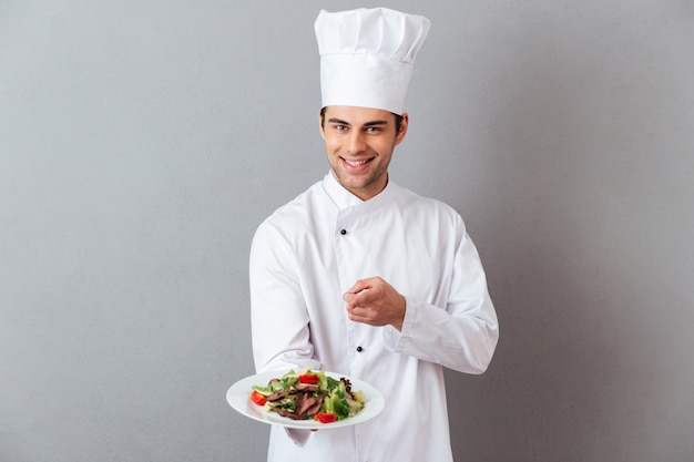 Foto gratuita alegre joven cocinero en uniforme sosteniendo ensalada apuntando a usted.