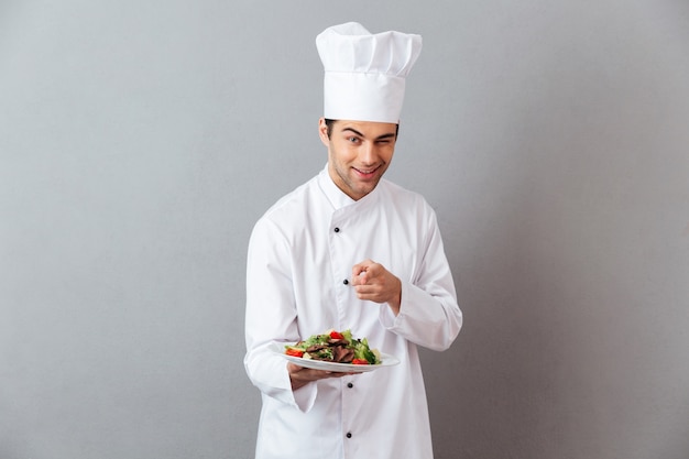 Alegre joven cocinero en uniforme sosteniendo ensalada apuntando a usted.