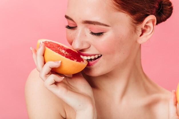Alegre joven comiendo pomelo. Dama de jengibre sonriente disfrutando de cítricos sobre fondo rosa.