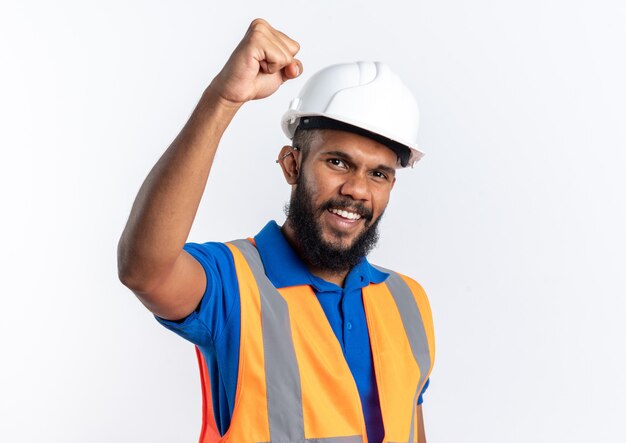 Alegre joven constructor hombre en uniforme con casco de seguridad de pie con el puño levantado aislado en la pared blanca con espacio de copia