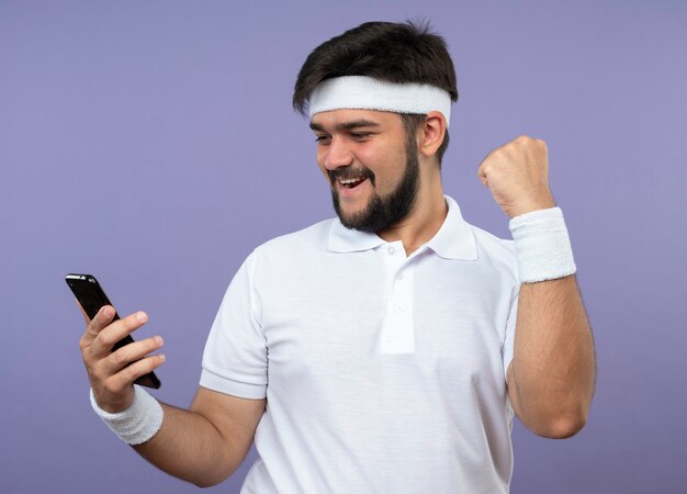 Alegre joven deportivo con diadema y muñequera sosteniendo y mirando el teléfono mostrando sí gesto