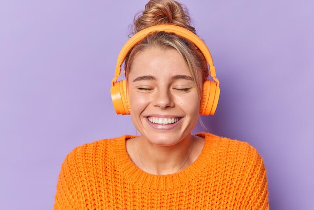 Alegre joven despreocupada sonríe con los dientes mantiene los ojos cerrados vestida con un jersey de punto cálido escucha su pista de audio favorita a través de auriculares posa en el interior sobre un fondo púrpura. Concepto de emociones felices