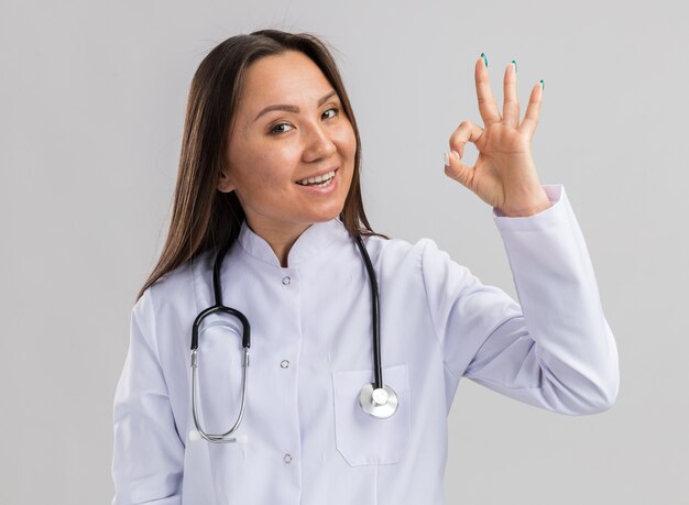 Alegre joven doctora asiática vistiendo bata médica y un estetoscopio mirando a la cámara haciendo el signo de ok aislado en la pared blanca