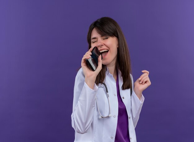 Alegre joven doctora en bata médica con estetoscopio pretende cantar con teléfono sobre fondo violeta aislado con espacio de copia