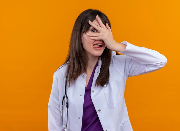 Alegre joven doctora en bata médica con estetoscopio saca la lengua y mira a través de los dedos de su mano sobre fondo naranja aislado con espacio de copia