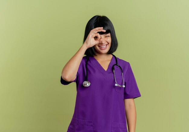 Alegre joven doctora morena en uniforme con estetoscopio mira a través de los dedos aislados sobre fondo verde oliva con espacio de copia