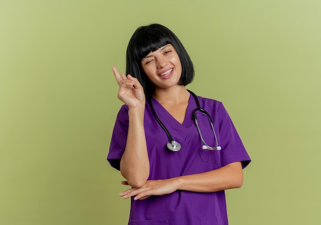 Alegre joven doctora morena en uniforme con estetoscopio parpadea y apunta hacia arriba