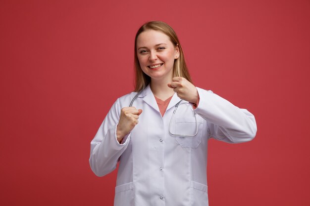 Alegre joven doctora rubia vistiendo bata médica y un estetoscopio alrededor del cuello haciendo gesto de boxeo