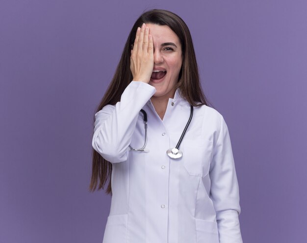 Alegre joven doctora vistiendo bata médica con estetoscopio cubre el ojo con la mano en púrpura