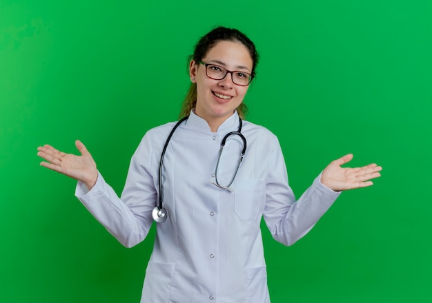 Alegre joven doctora vistiendo bata médica y estetoscopio y gafas mostrando las manos vacías aisladas en la pared verde