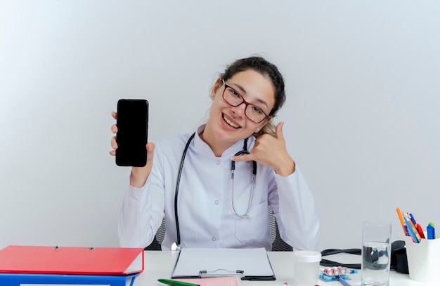 Alegre joven doctora vistiendo bata médica y estetoscopio y gafas sentado en el escritorio con herramientas médicas mostrando teléfono móvil mirando haciendo gesto de llamada aislado