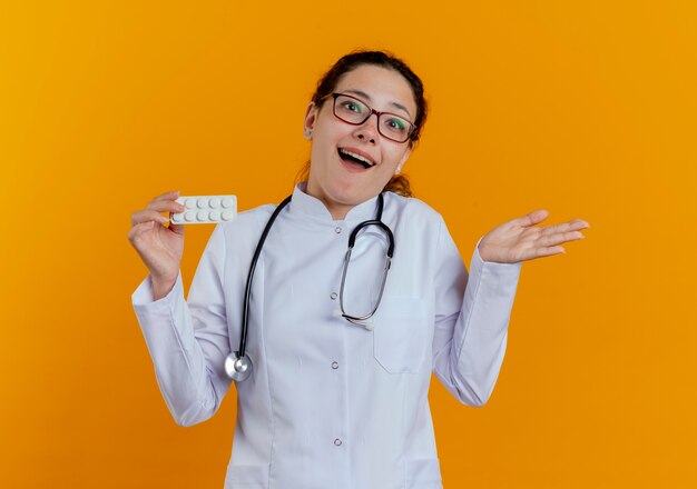 Alegre joven doctora vistiendo bata médica y estetoscopio con gafas sosteniendo pastillas y extendiendo las manos aisladas